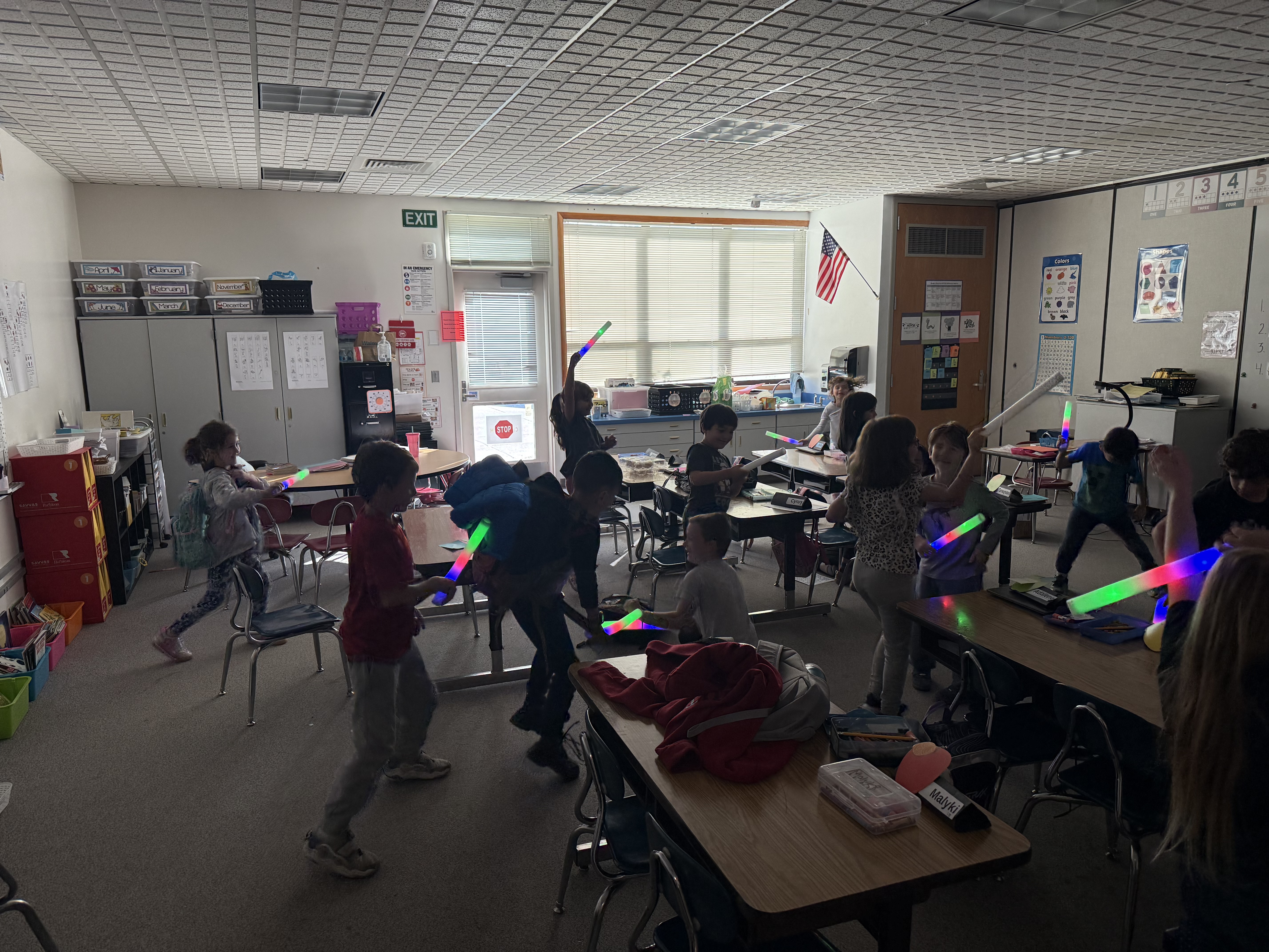 A group of students excitedly play with colorful light-up foam sticks in a brightly lit classroom.