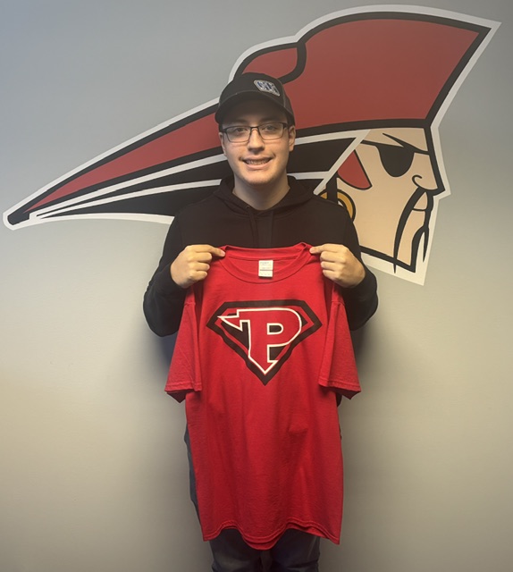 A smiling young man wearing a black hoodie, glasses, and a cap holds up a red t-shirt with a white and black 'P' logo in front of a pirate mascot wall graphic.