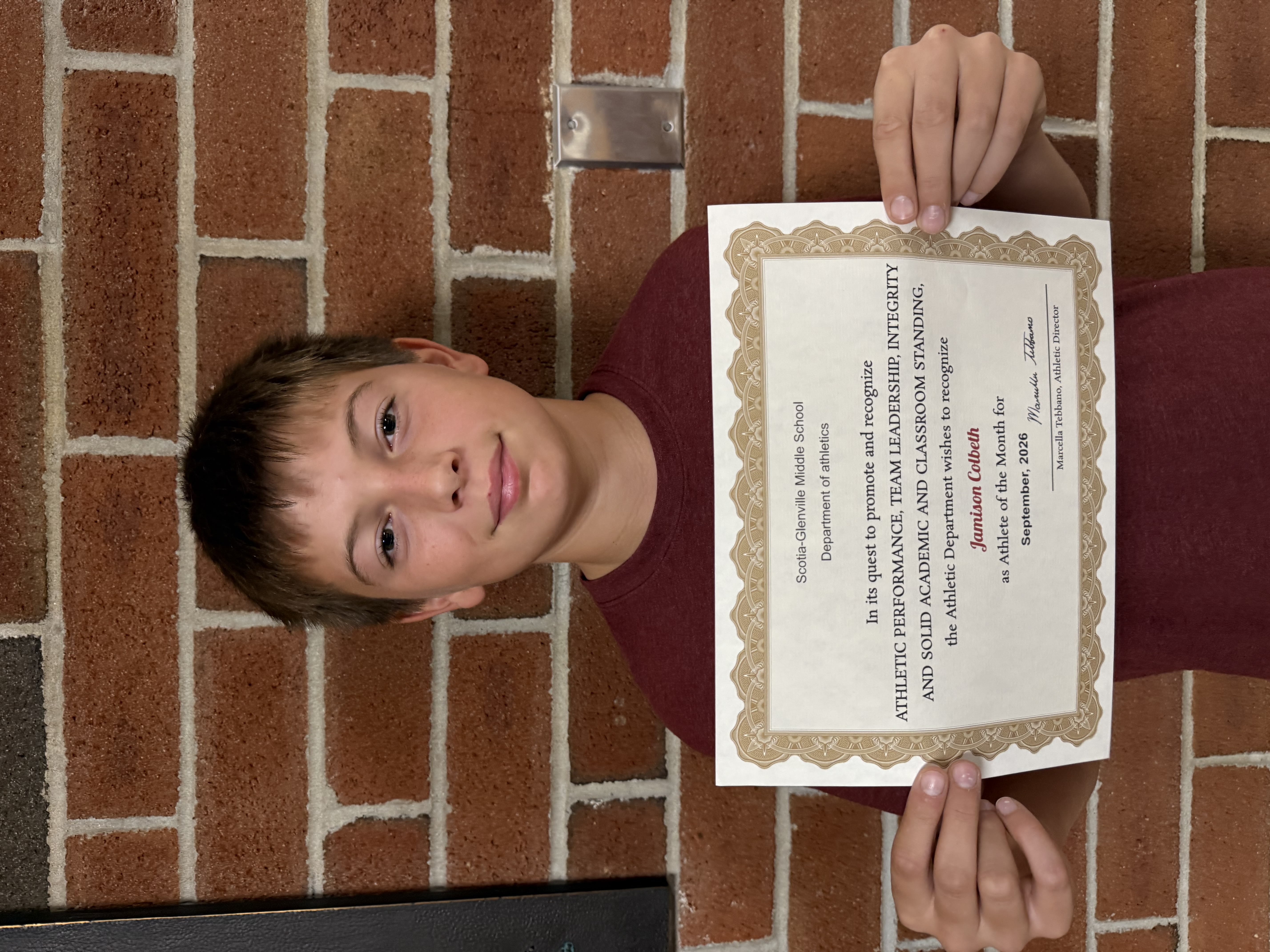 A young person stands in front of a brick wall, proudly holding up an award certificate.