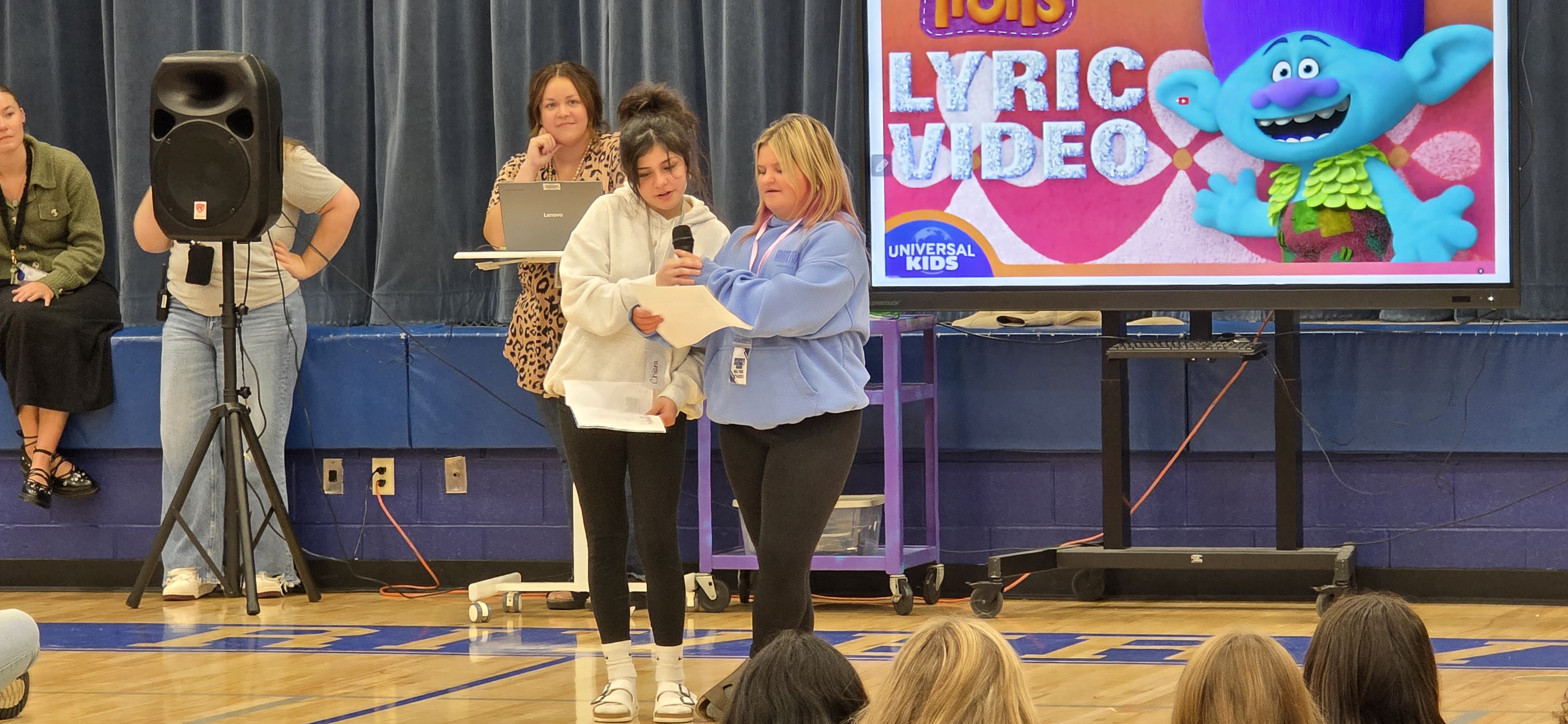 Two girls in hoodies read from papers while holding a microphone on stage in a school gymnasium.