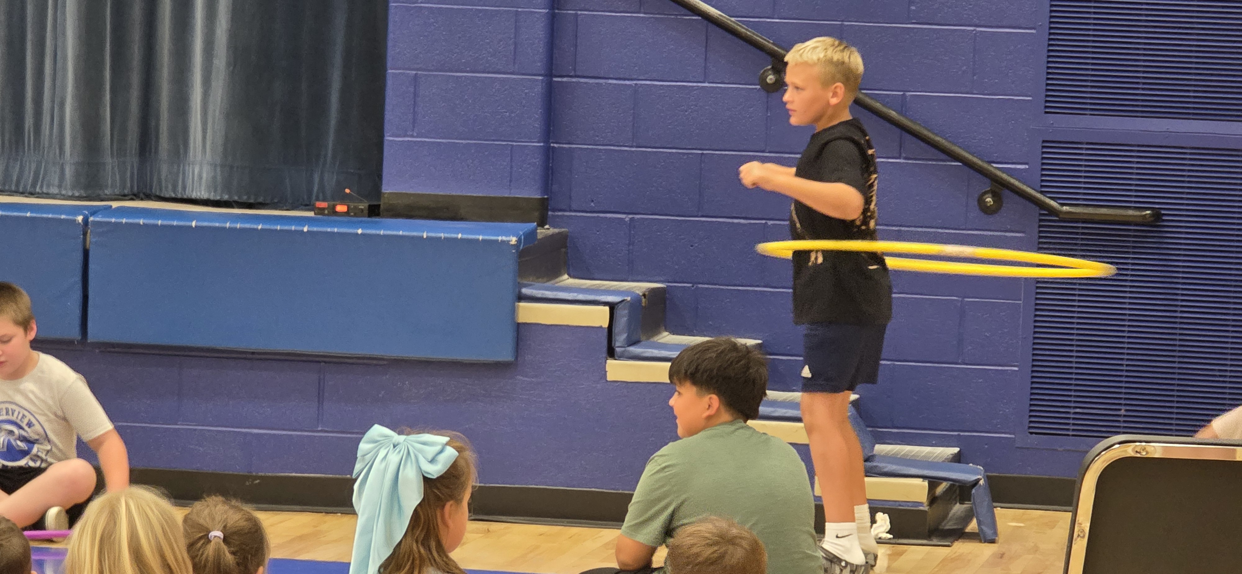 A young boy hula hoops on gym steps while classmates watch in an indoor gymnasium.