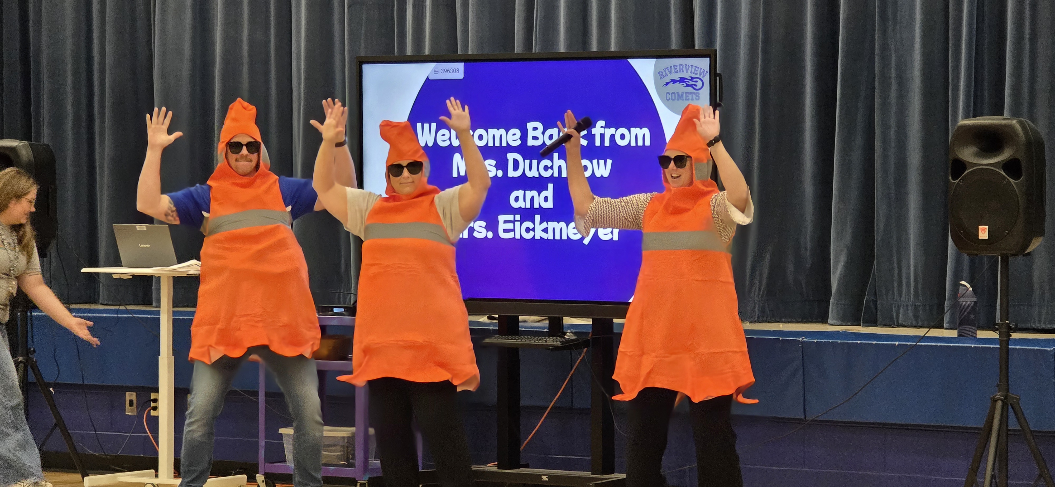 Three people dressed as orange traffic cones with sunglasses wave enthusiastically on a stage.