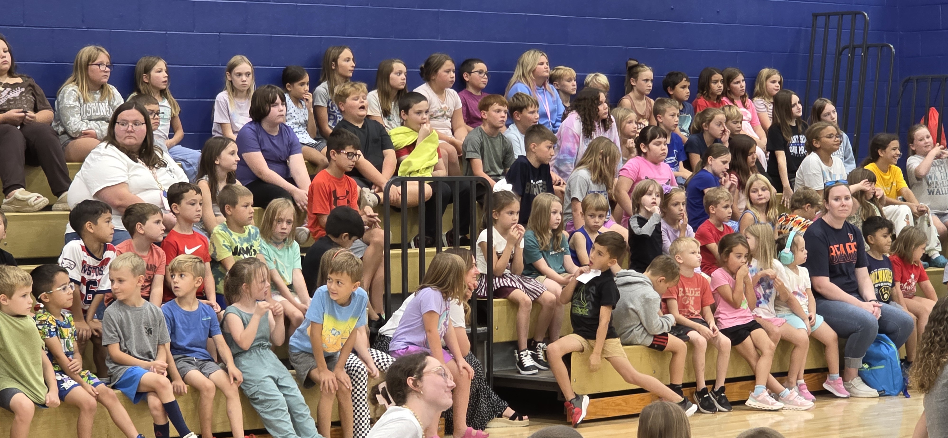 A large group of children and adults sit on wooden bleachers in a gymnasium.