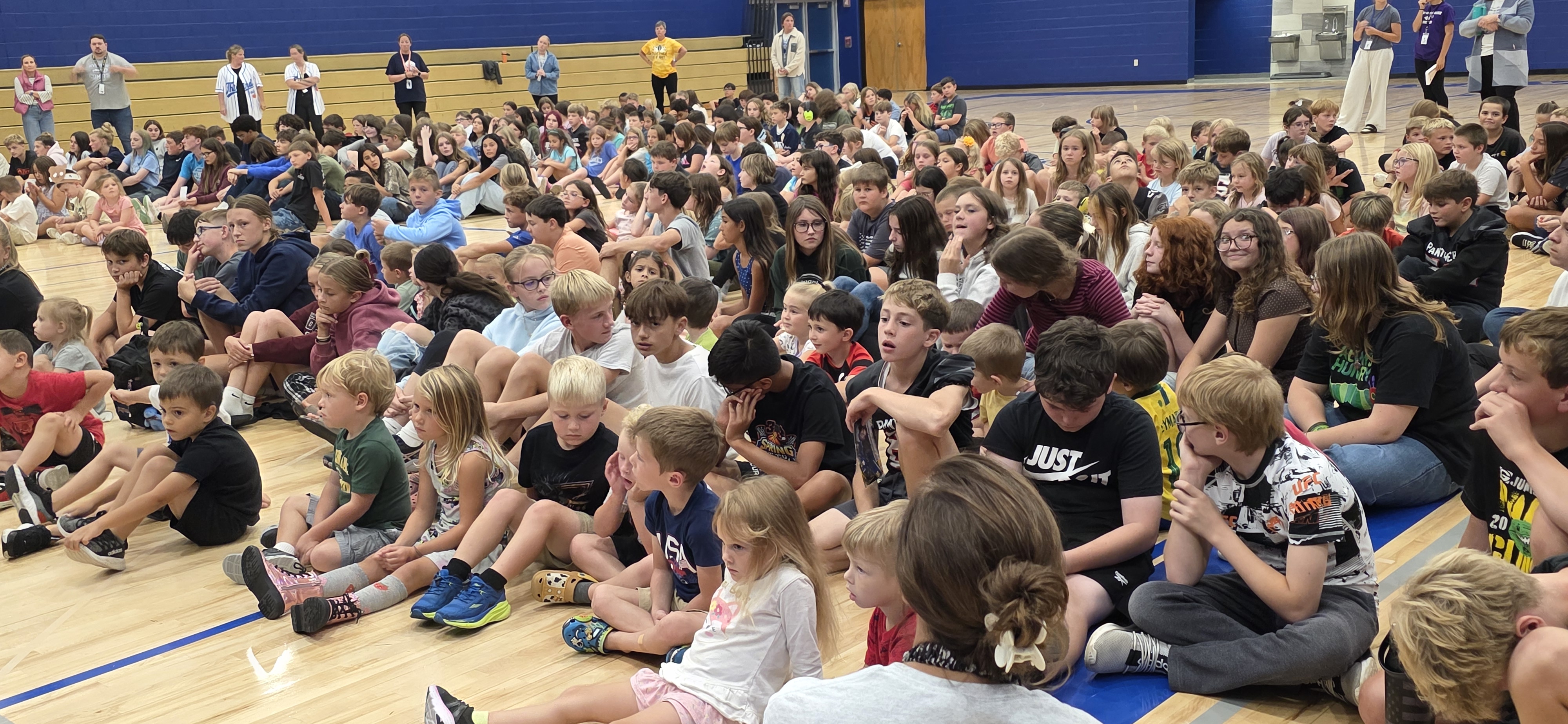 A large group of students sits attentively on the wooden floor of a school gymnasium.