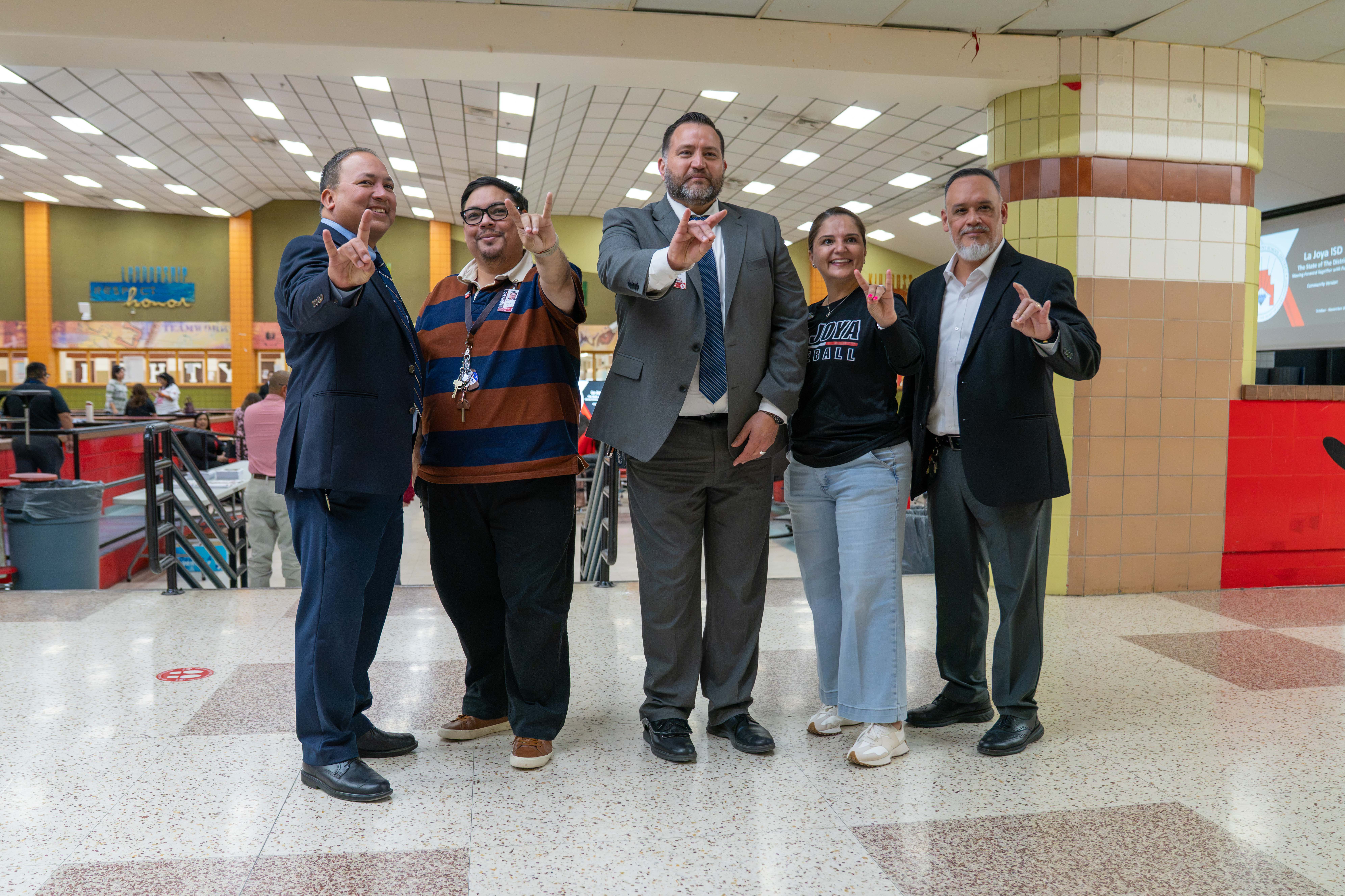 Five adults stand in a school hallway, smiling and holding up a hand sign together.