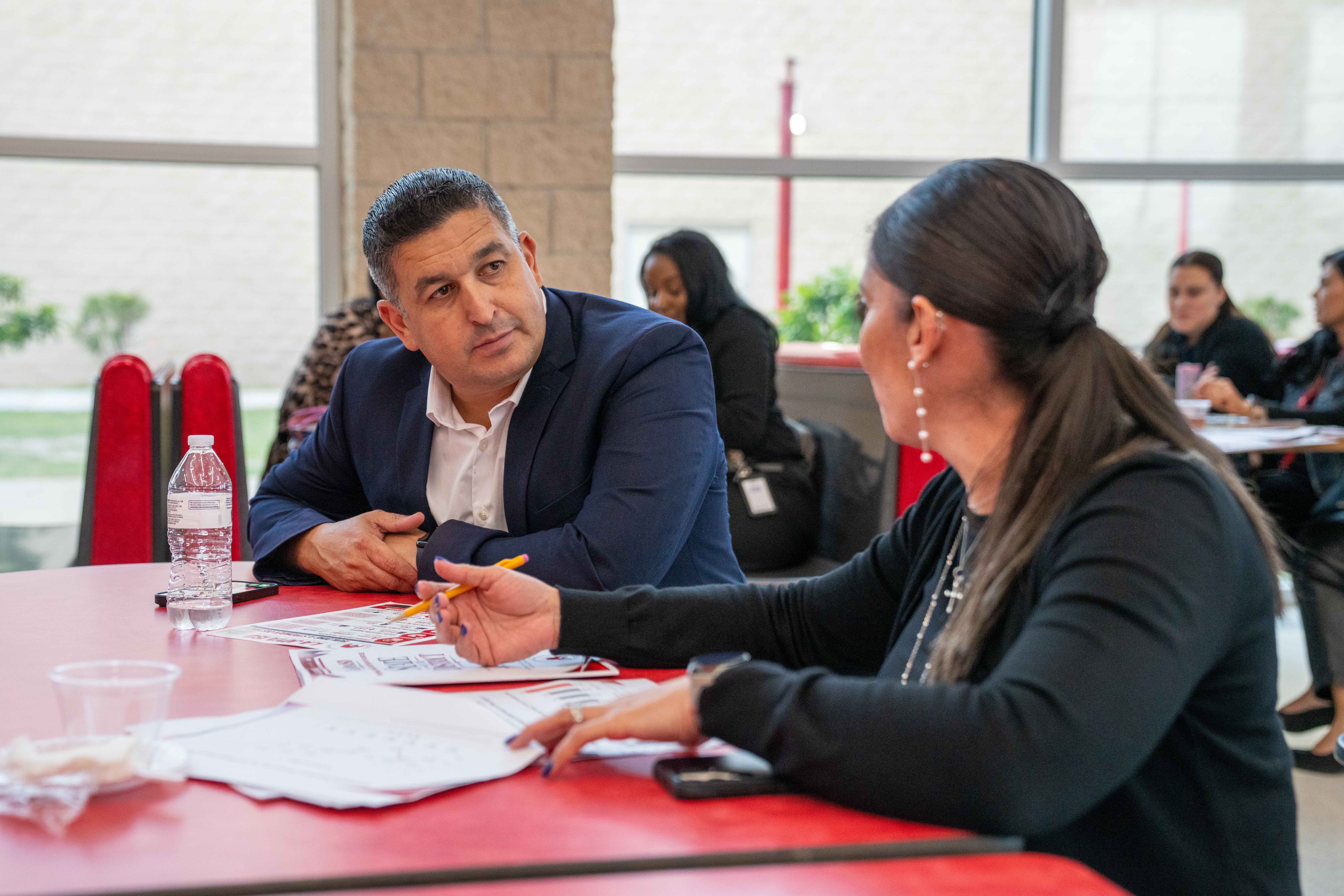 A man in a blue suit and white shirt listens intently to a woman speaking across a red table with documents.