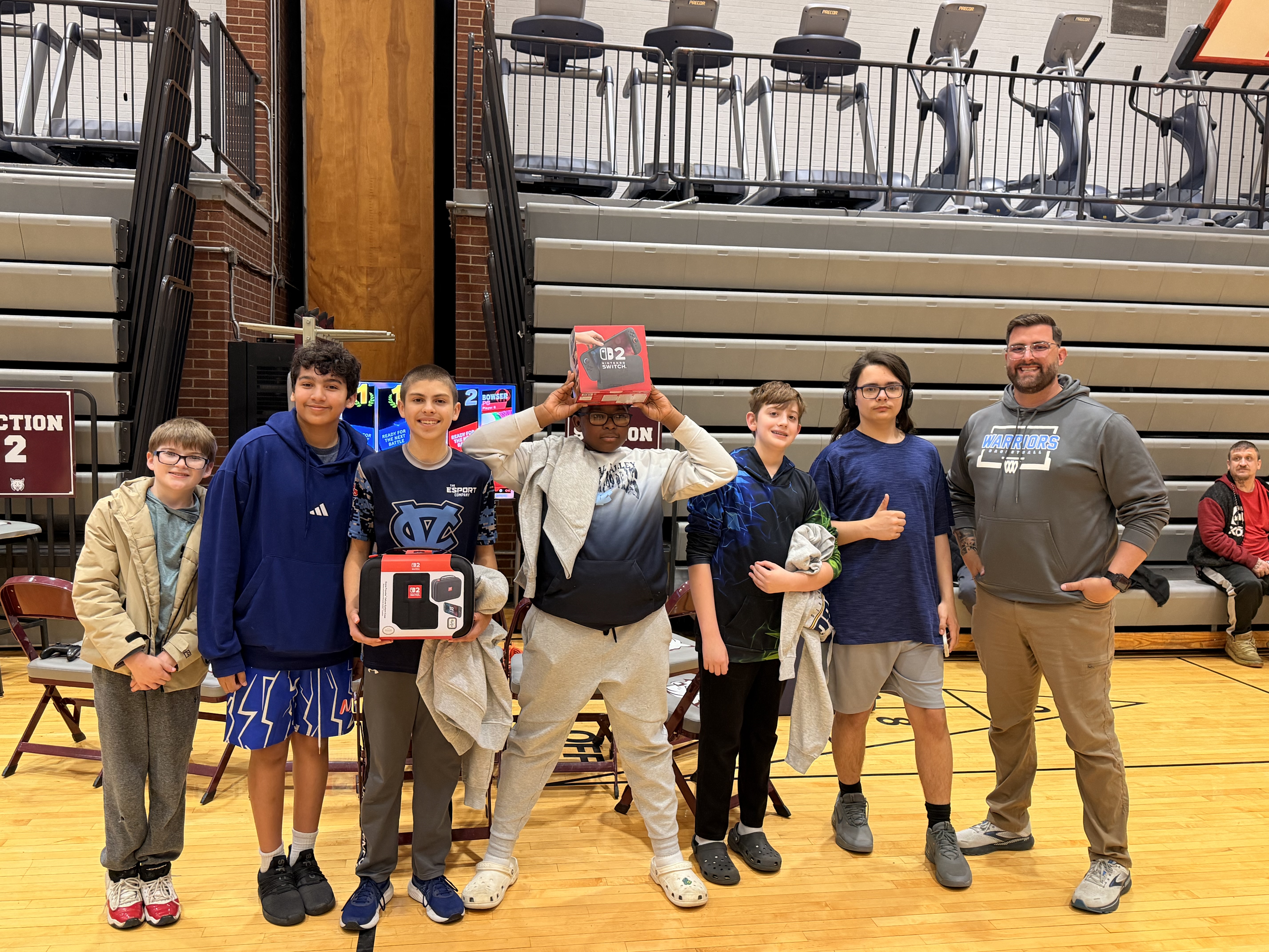 A group of students and an adult stand on a basketball court, holding Nintendo Switch boxes and accessories.