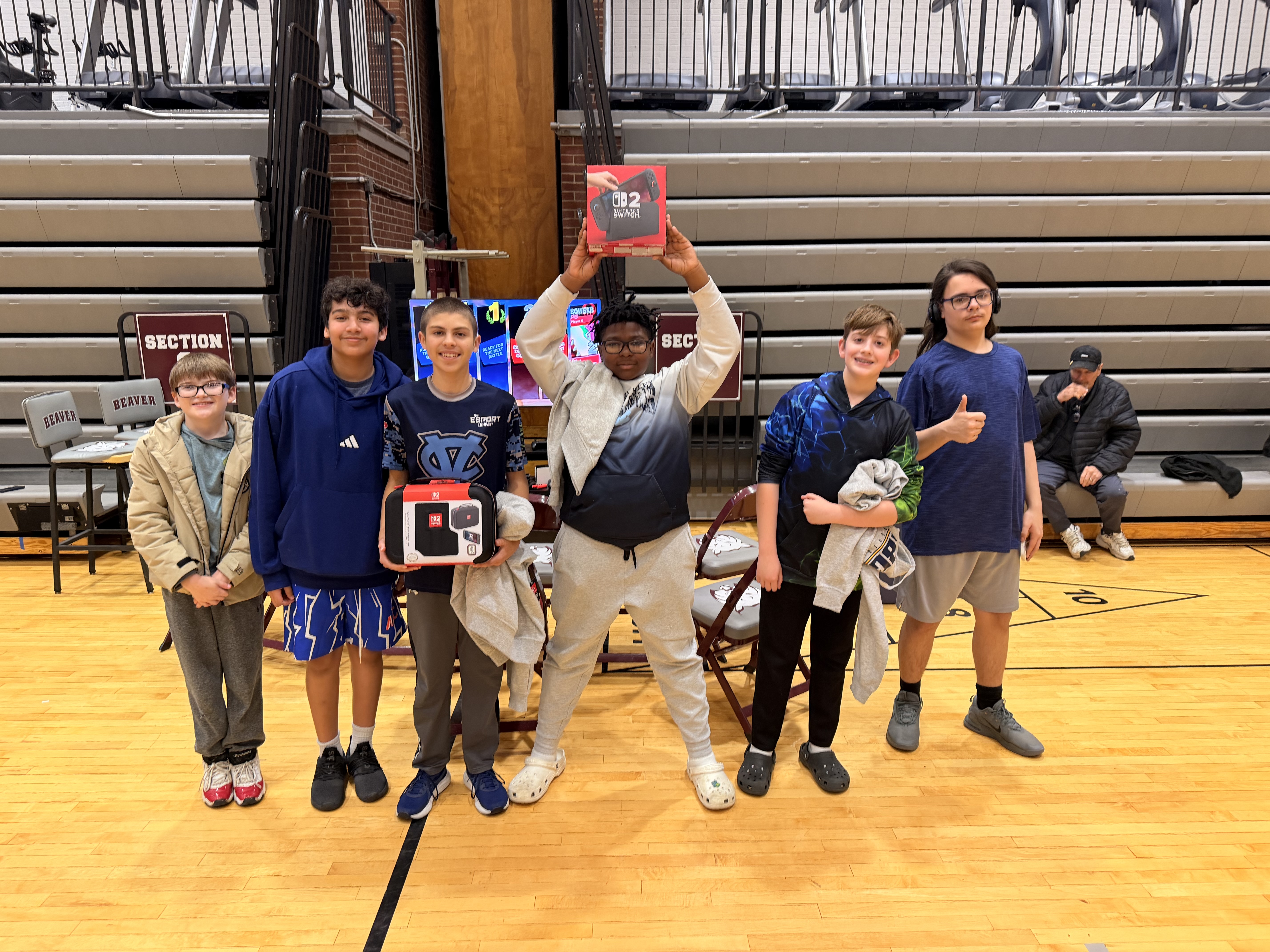 A group of young people stand on a wooden floor in a gymnasium, holding a Nintendo Switch box.