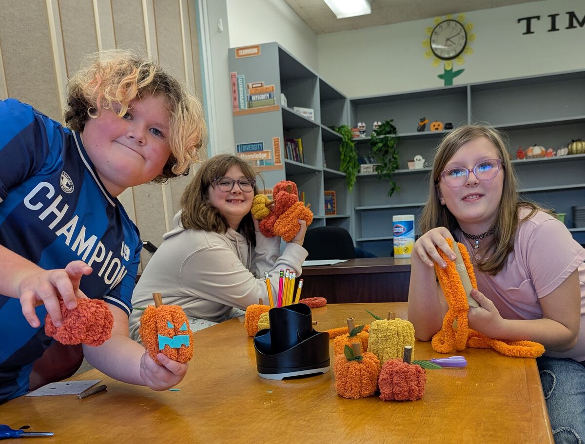Three children sit around a wooden table in a classroom, crafting small pumpkins out of orange and yellow yarn.