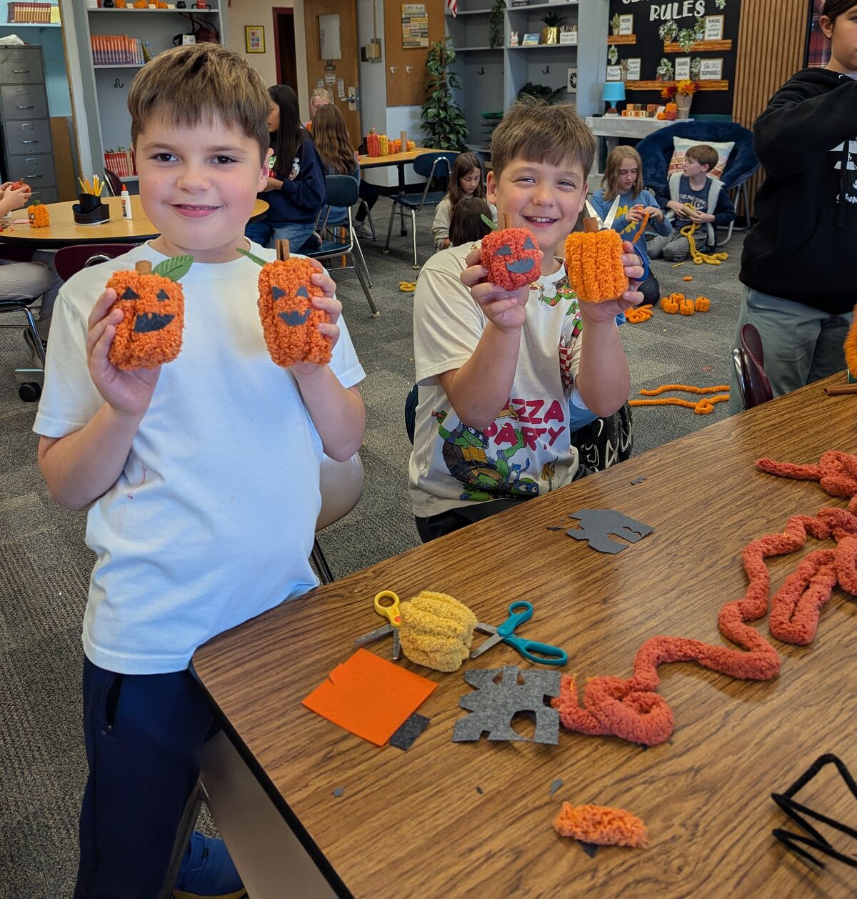 Two boys smile proudly as they show off yarn-made miniature pumpkins in a classroom crafting session.