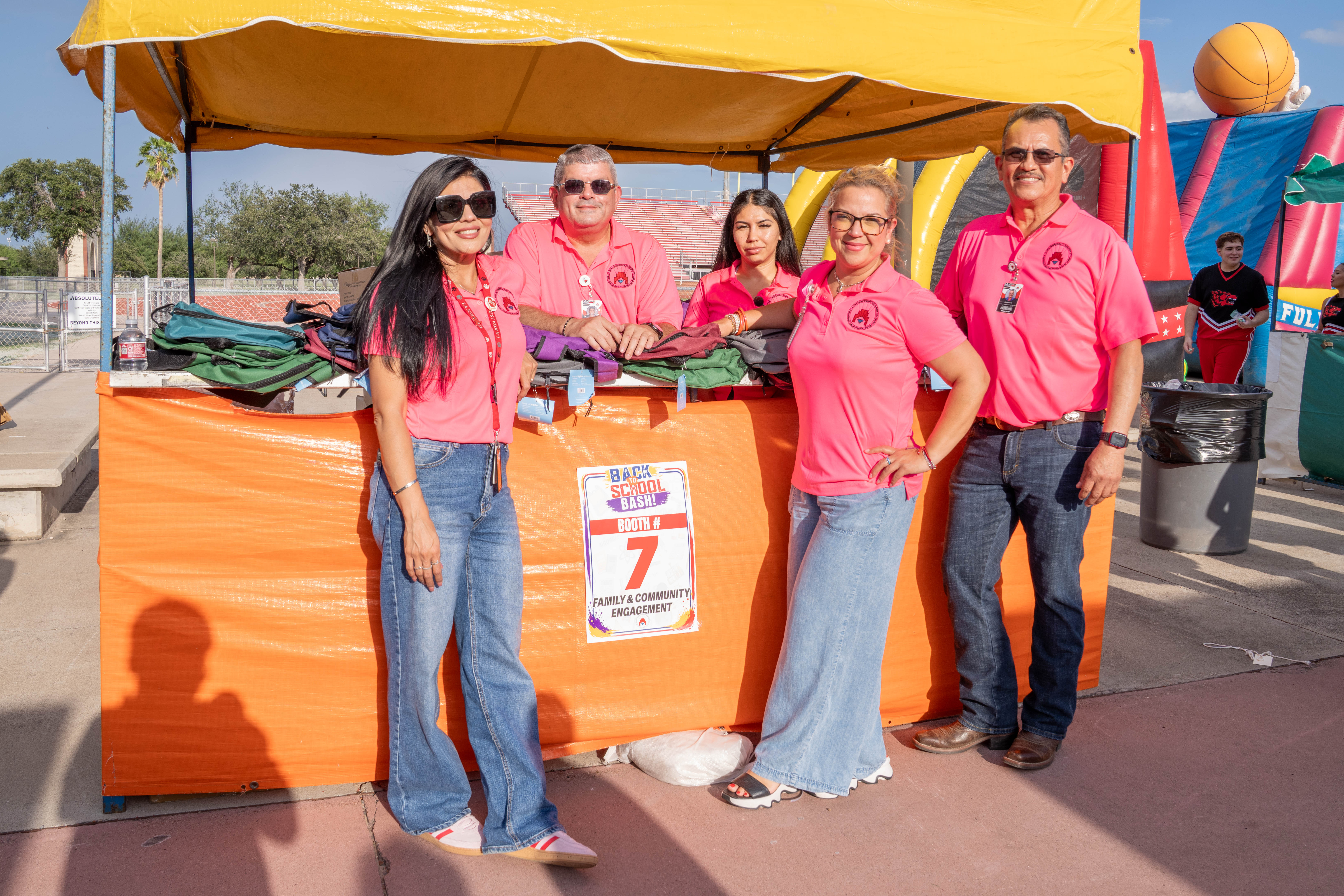 Five people in pink shirts stand behind an orange booth at a 'Back to School Bash' event.