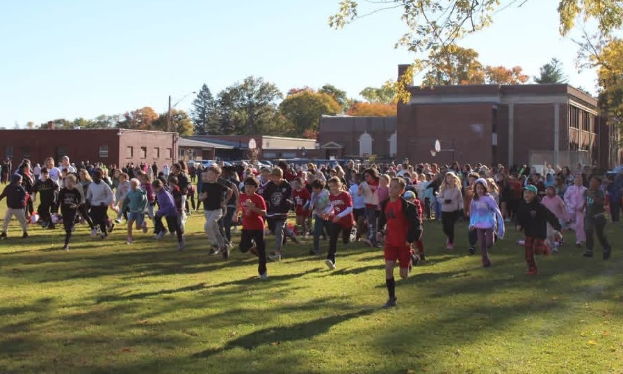 A large group of children participate in a fun run across a grassy field near school buildings.