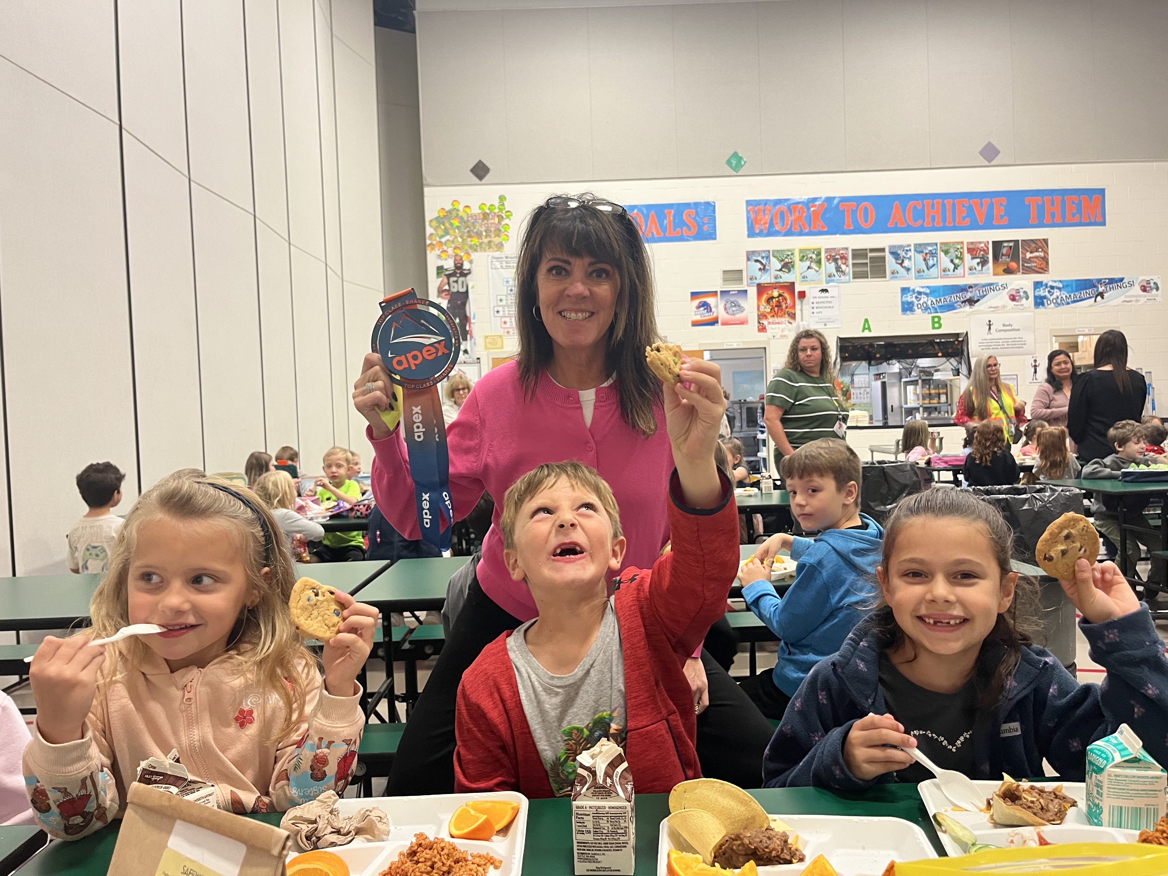 A smiling woman holds up an Apex medal while children happily show off cookies at a school lunch table.