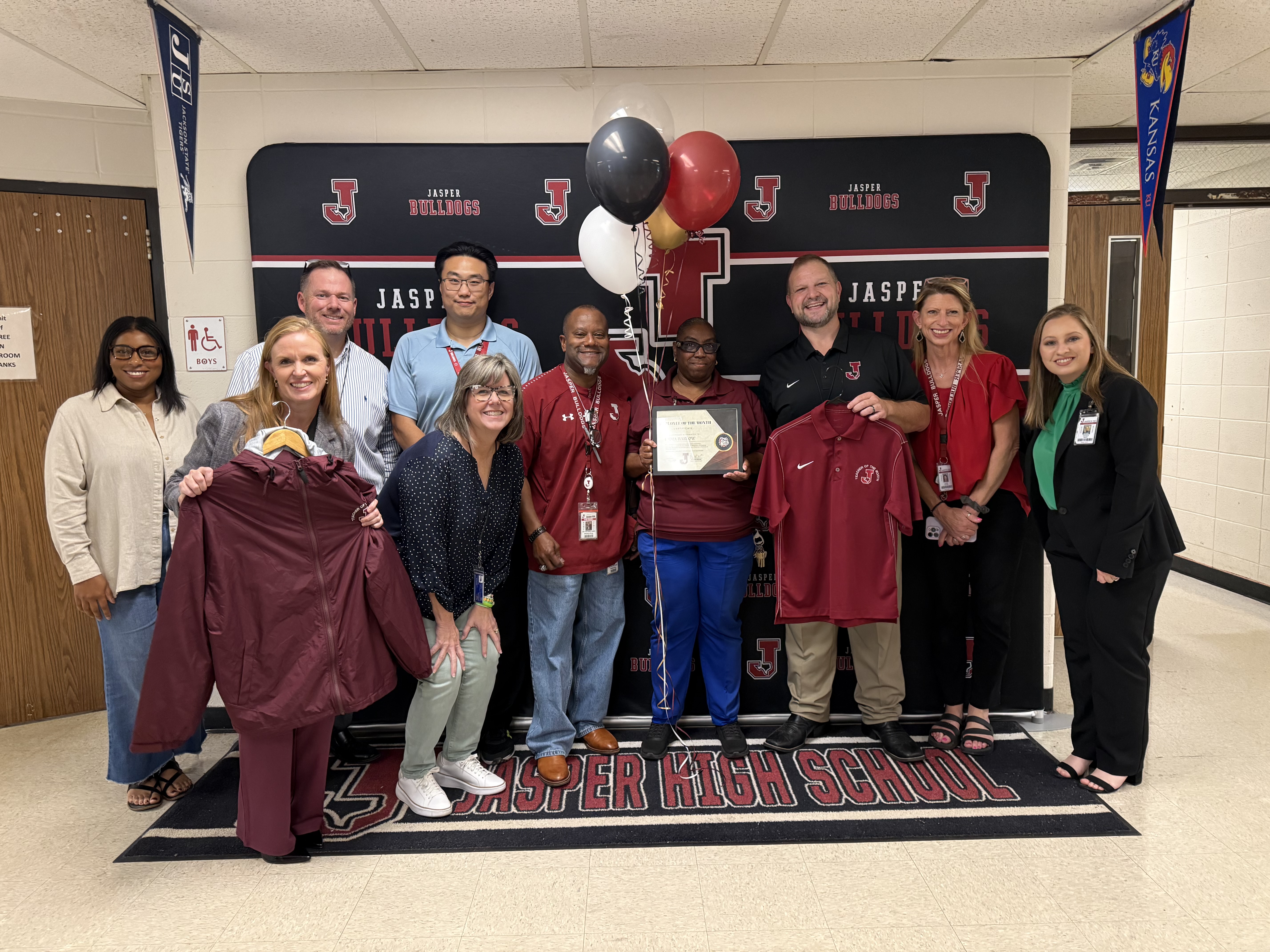 A diverse group of school staff members pose smiling for a photo to recognize Emma Barlow, Employee of the Month, in front of a Jasper Bulldogs banner holding awards and apparel.