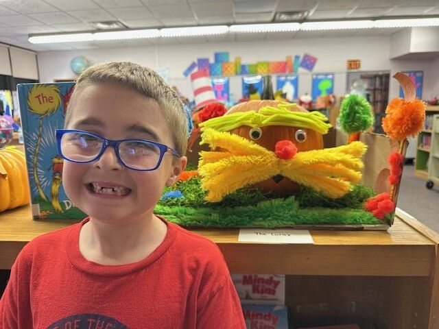 A smiling child with blue glasses and a red shirt stands next to a Lorax-themed pumpkin display in a classroom.