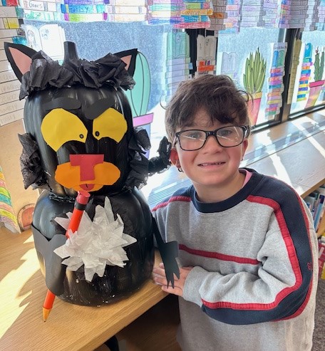 A smiling child poses proudly next to a large black pumpkin decorated like a cat with a pencil.