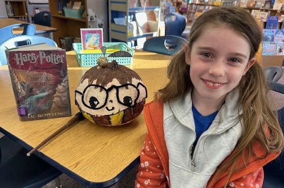 A smiling young girl sits in a library next to a Harry Potter book and a decorated Harry Potter pumpkin.