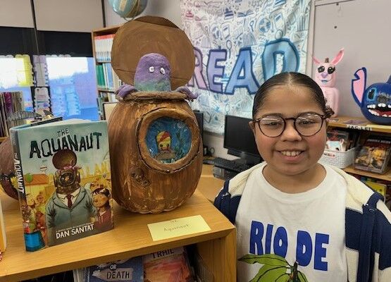 A smiling young person stands beside a book-themed craft project and the graphic novel The Aquanaut in a library.