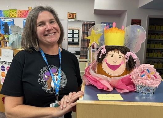 A smiling woman stands next to a decorated pumpkin dressed as a fairy princess with a crown and wings.