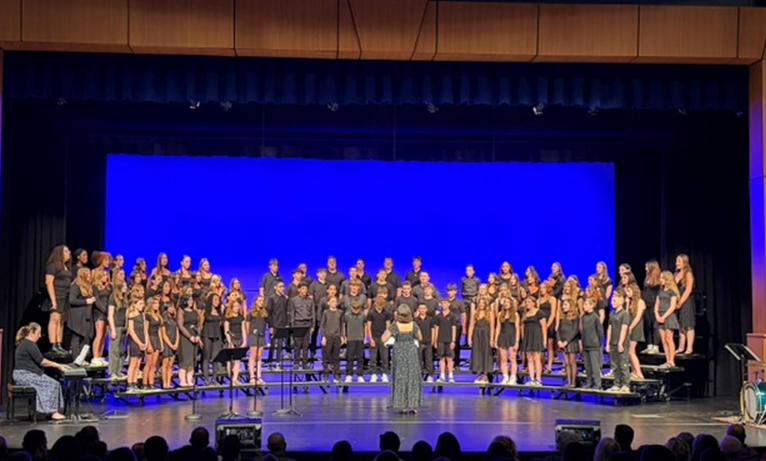 A large choir of students in black attire stands on risers on a stage, facing a conductor.