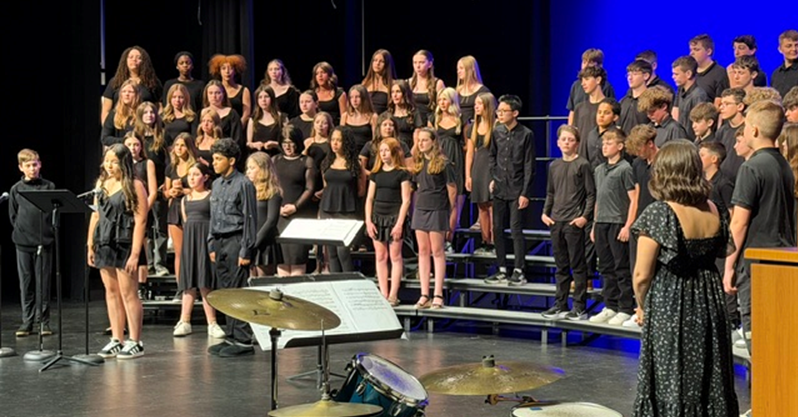 A large choir of students in black attire stands on risers on a stage, facing forward.