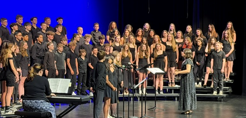 A choir of students in black attire stands on risers, facing a conductor and a pianist.