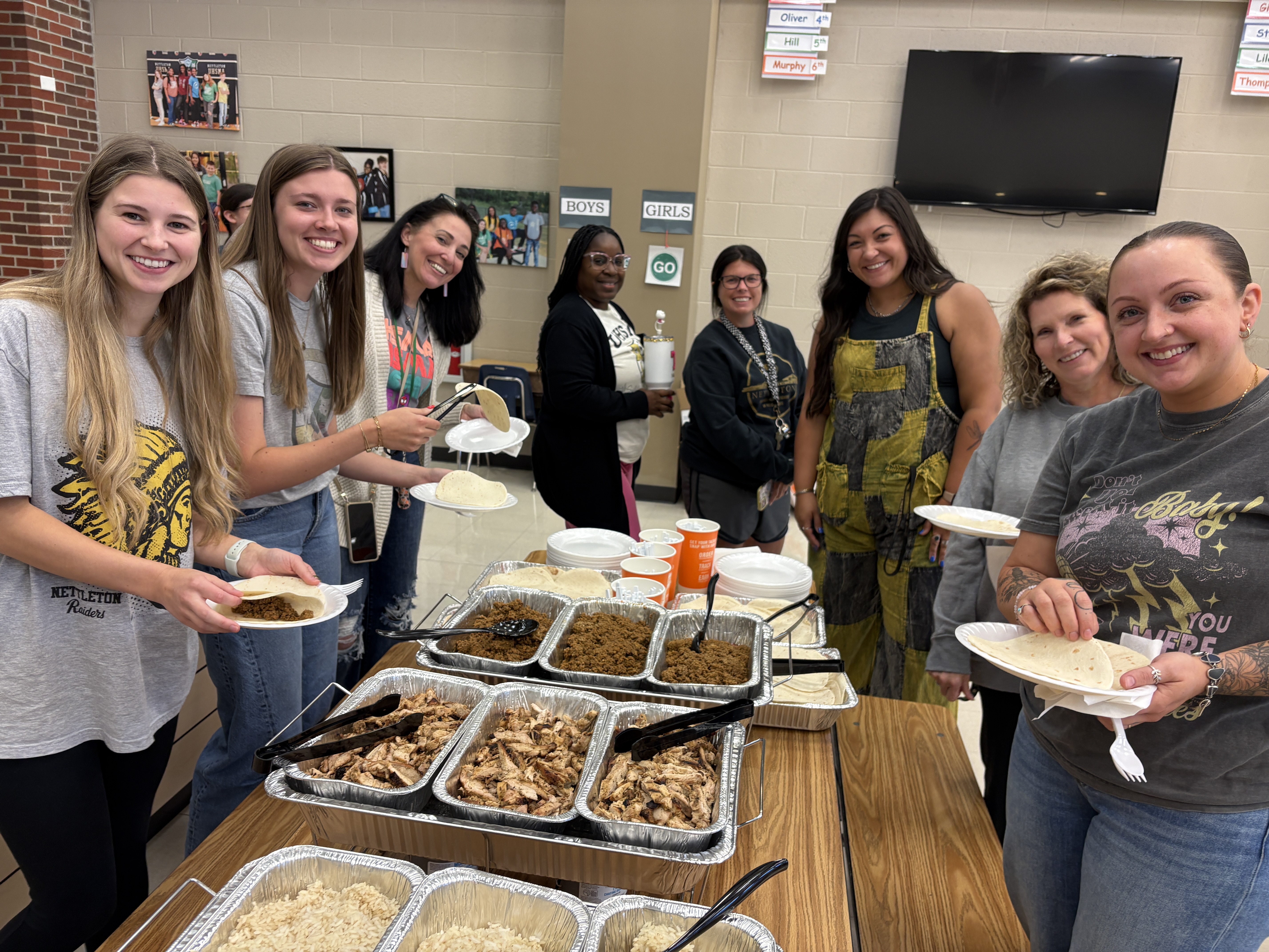 A group of smiling women are gathered around a table with a taco bar, preparing their meals.