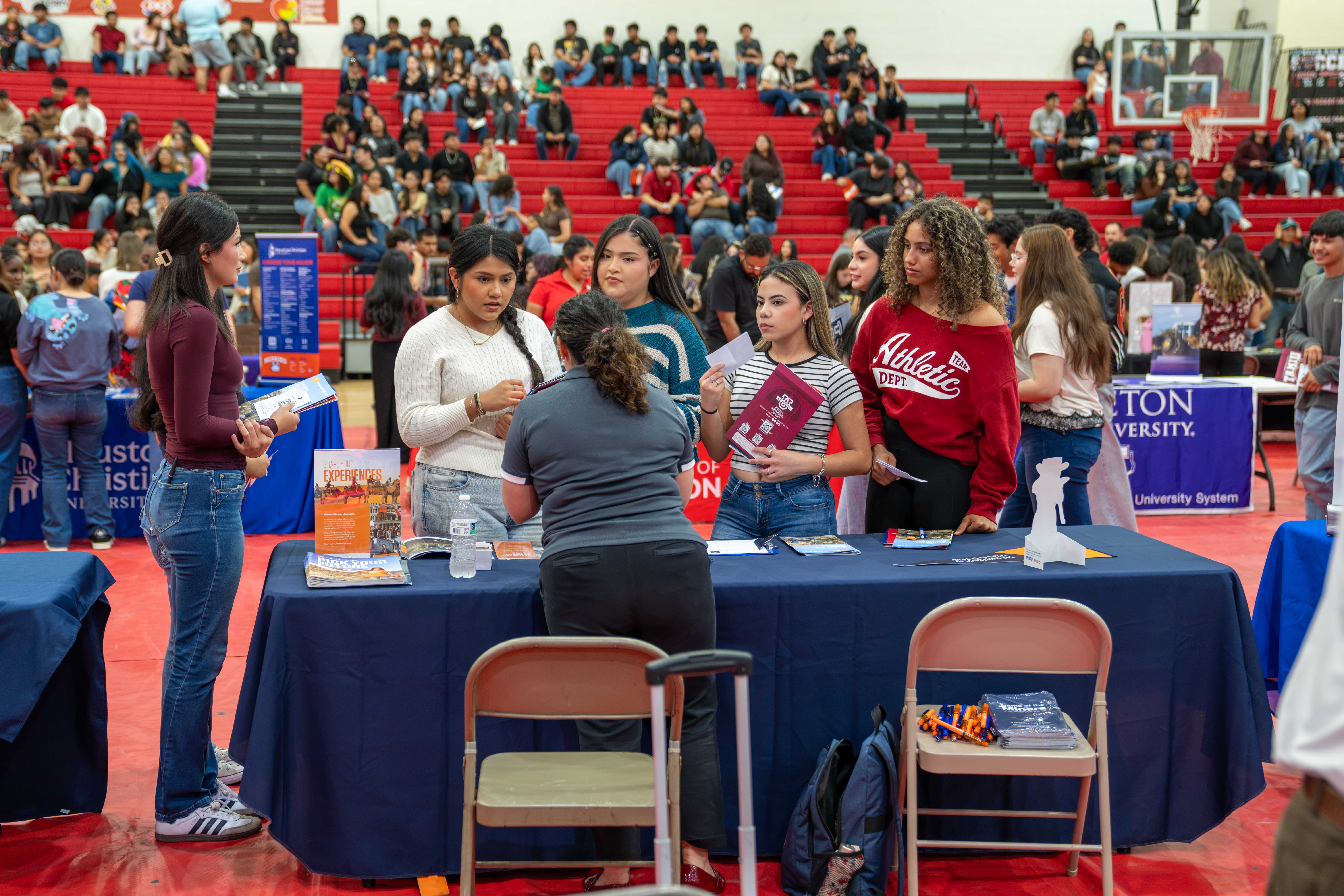 Students gather at a university career fair booth inside a gymnasium, looking at informational brochures.