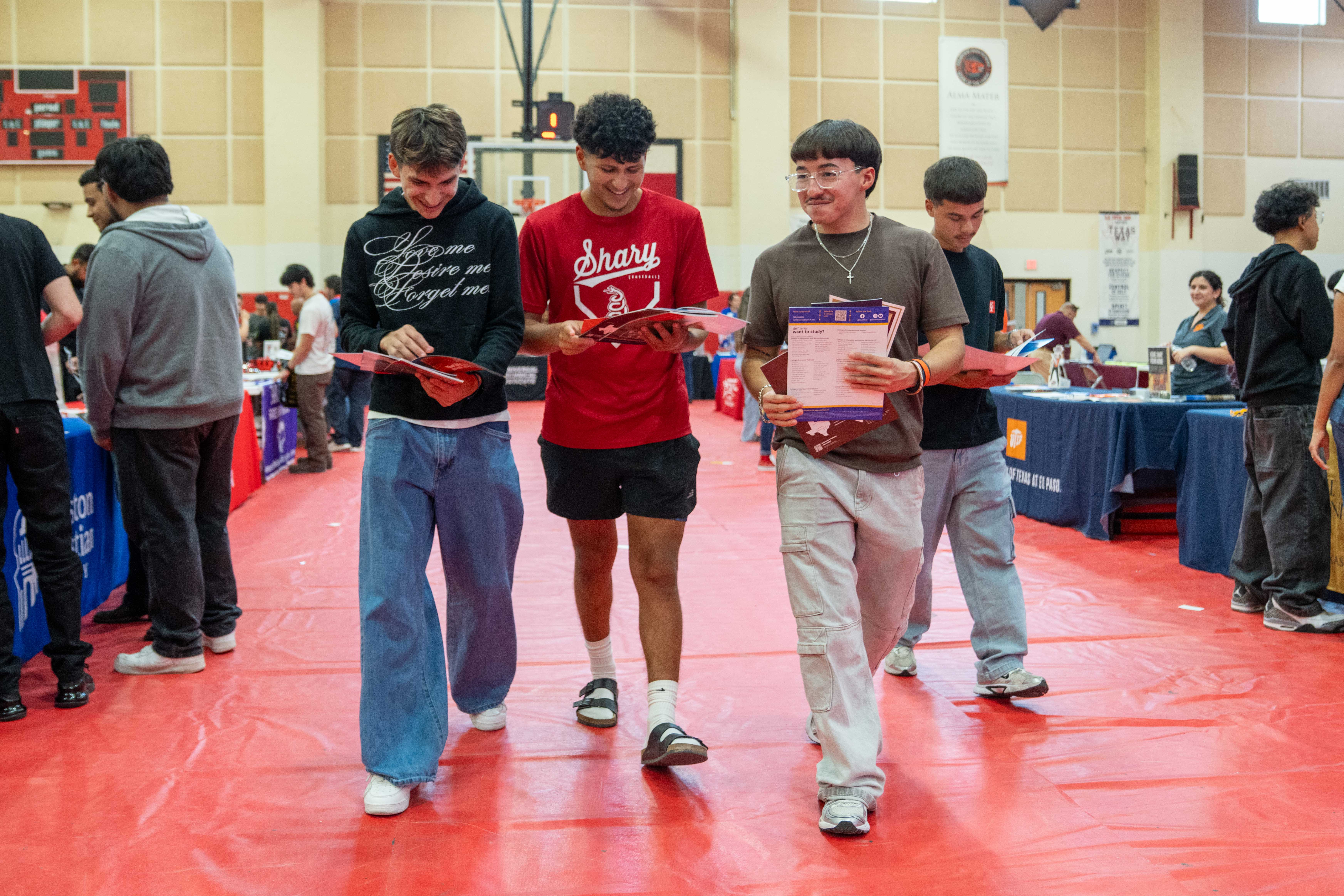 Three young men walk together across a red gym floor, smiling as they look at informational brochures during a school event.