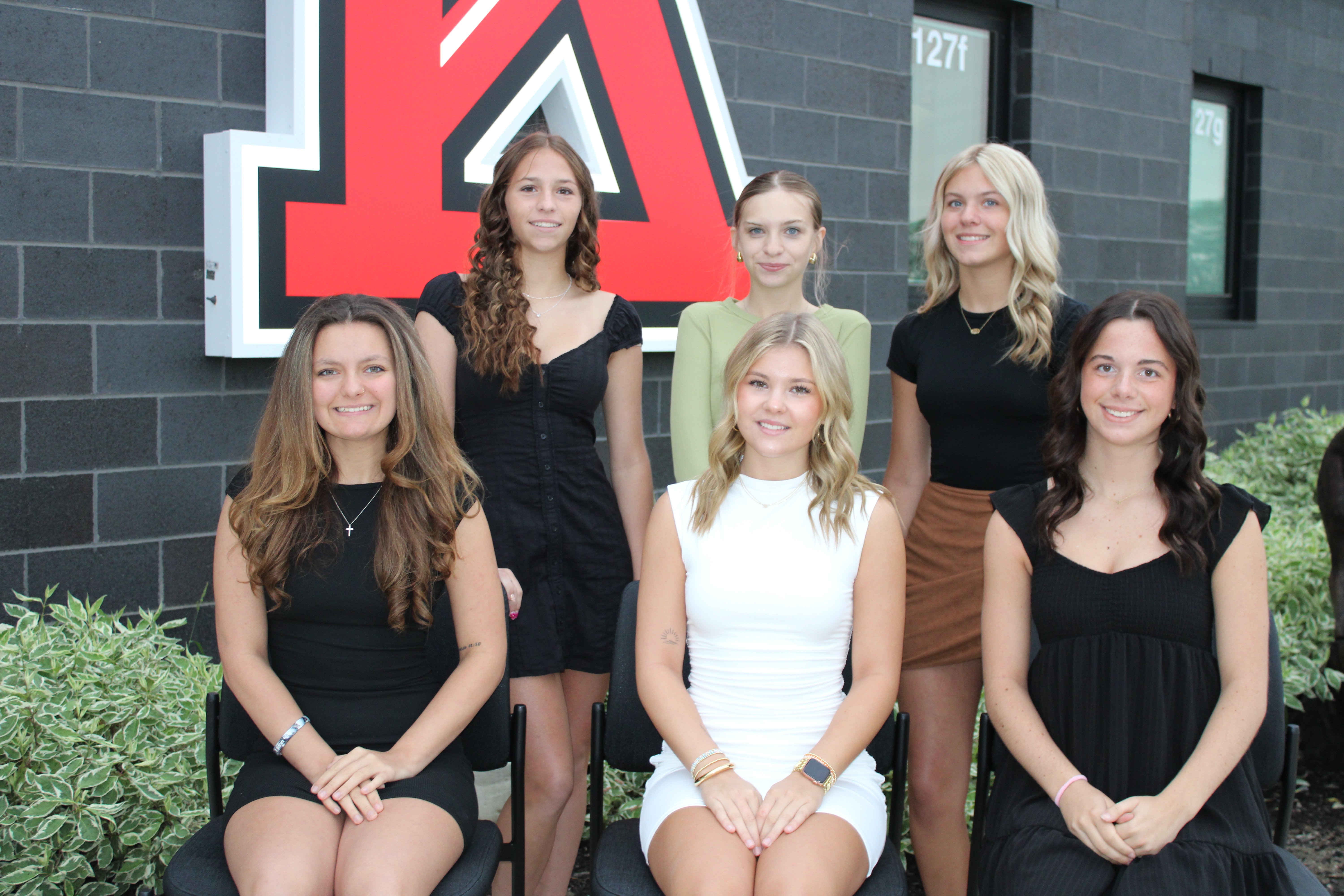 A group of six smiling young women pose together outdoors in front of a dark brick wall with a large red letter logo.