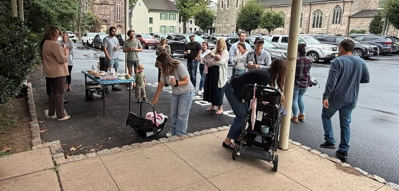 A group of people gather outdoors next to a parking lot for a community event near a food table.