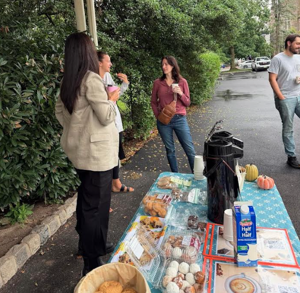 People gather around a table outdoors on a street, chatting near a display of baked goods and coffee dispensers.