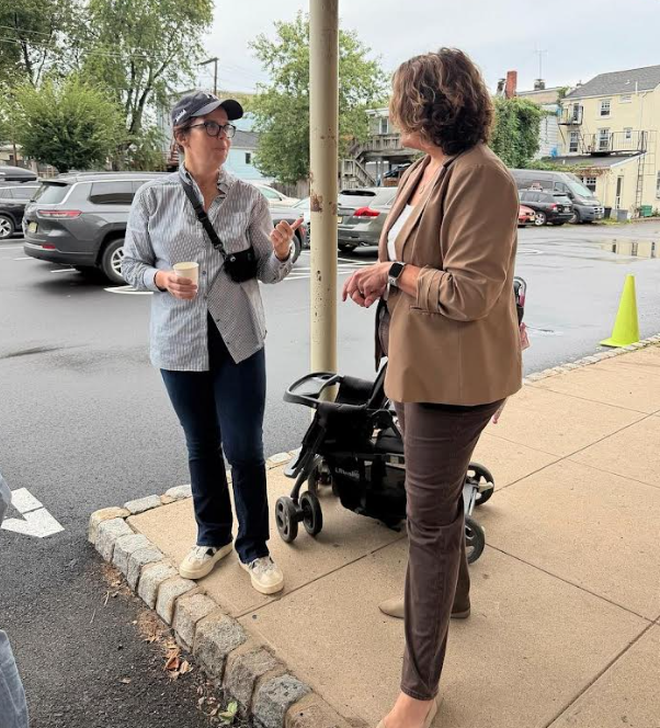 Two women stand on a sidewalk, engaged in a conversation near a parking lot.