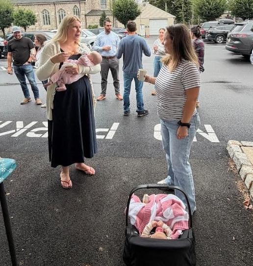 A group of people socialize in an asphalt parking lot, with two women chatting near a baby carrier.
