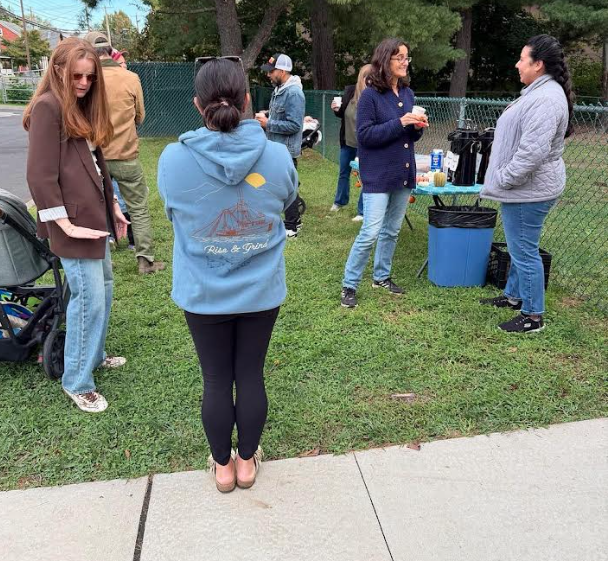 A group of people gather on a grassy area outdoors, chatting and enjoying coffee from a self-serve table setup.
