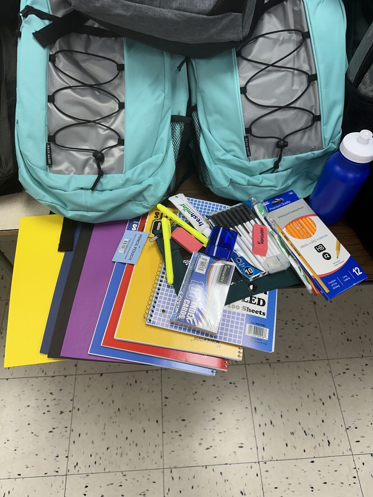 Two teal backpacks are packed with school supplies including notebooks, pencils, and a water bottle.