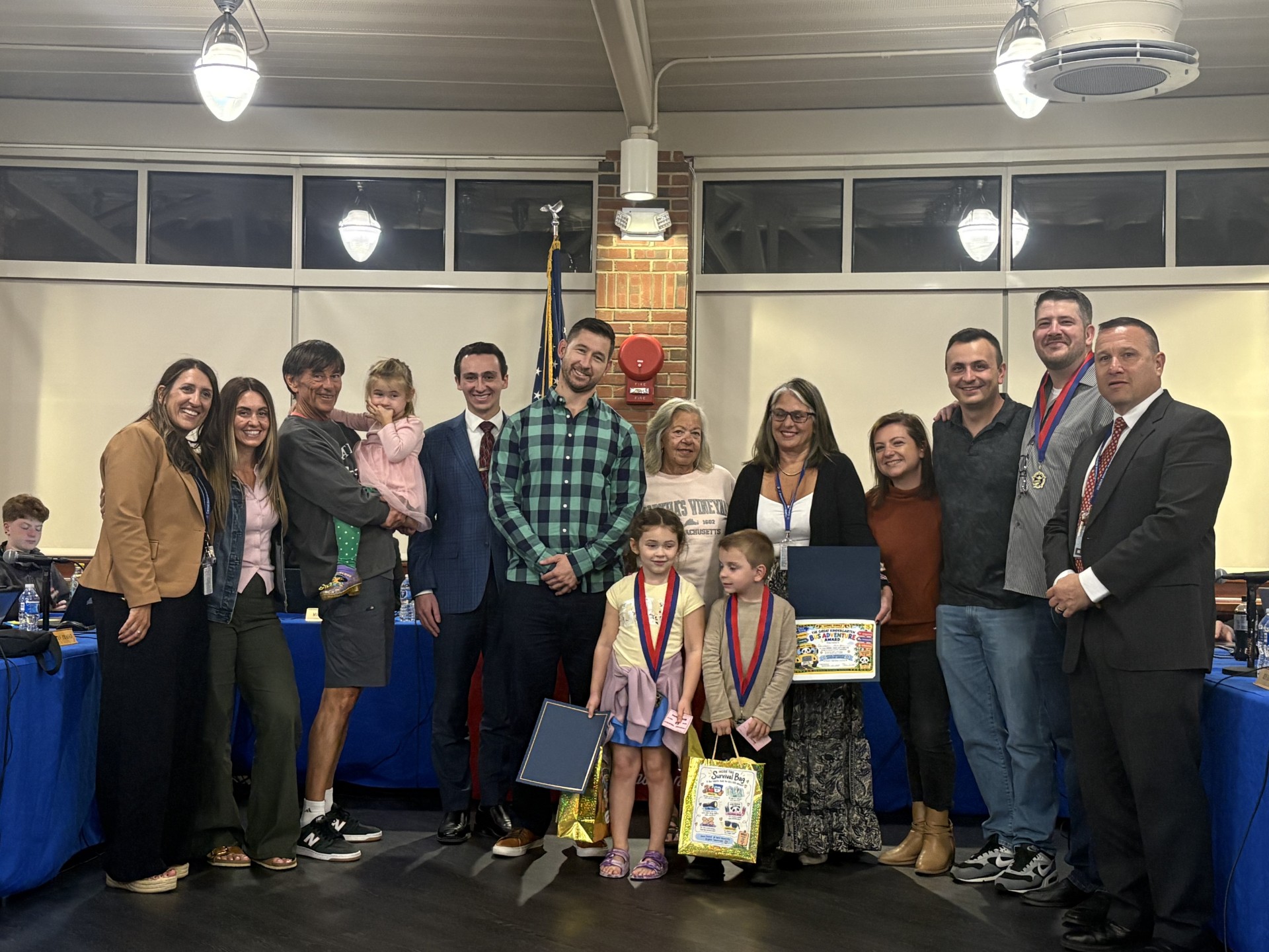 A group of adults and children pose together indoors, smiling while holding awards and gift bags.