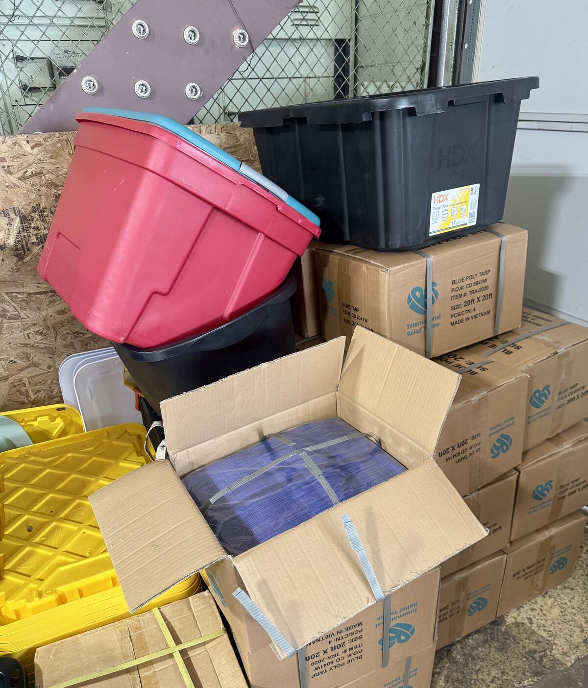 A stack of storage containers and boxes, including a red and blue tote, a black HDX tote, and several cardboard boxes labeled 'BLUE POLY TARP'.