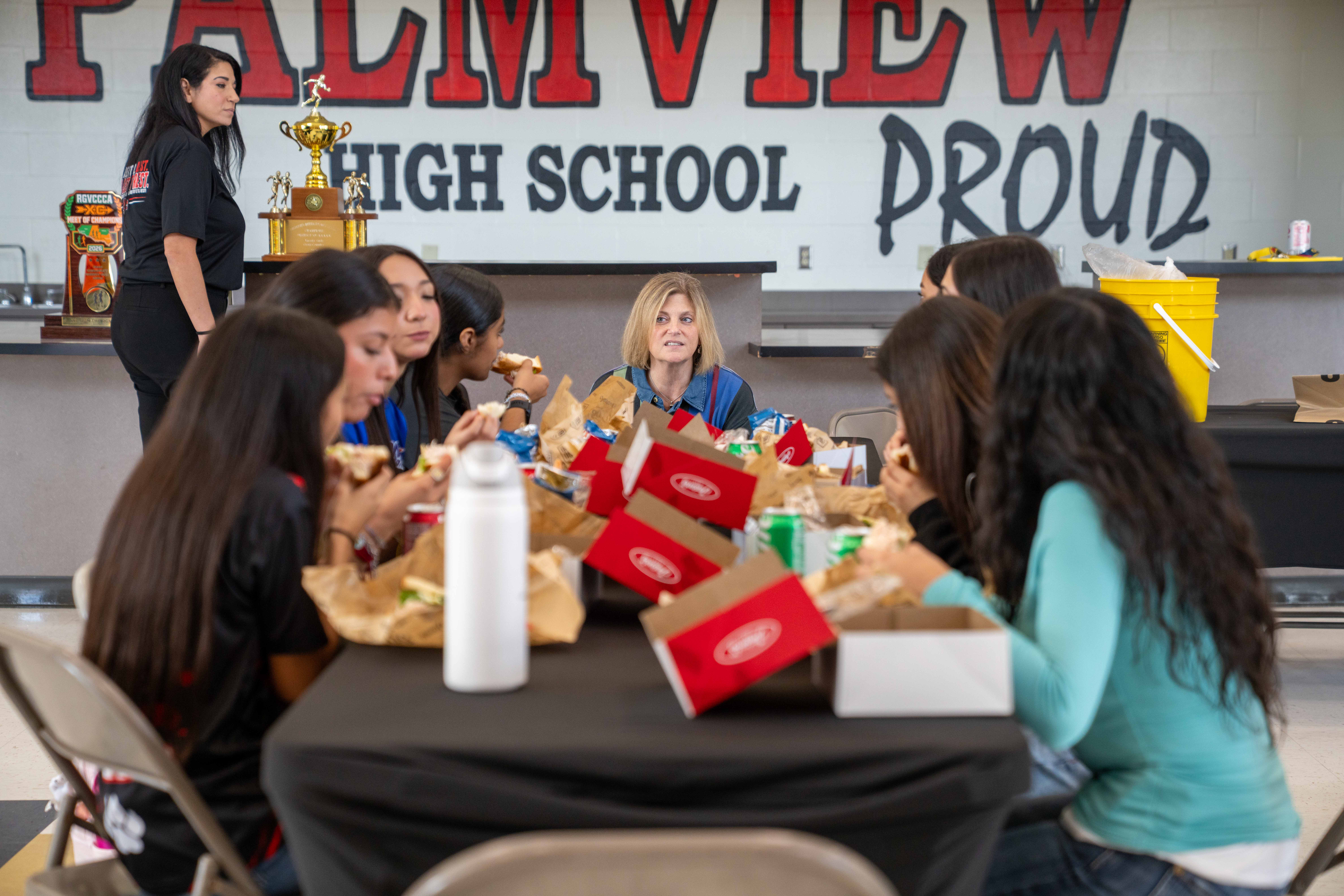 A group of students and teachers eat lunch together at tables in a high school cafeteria.