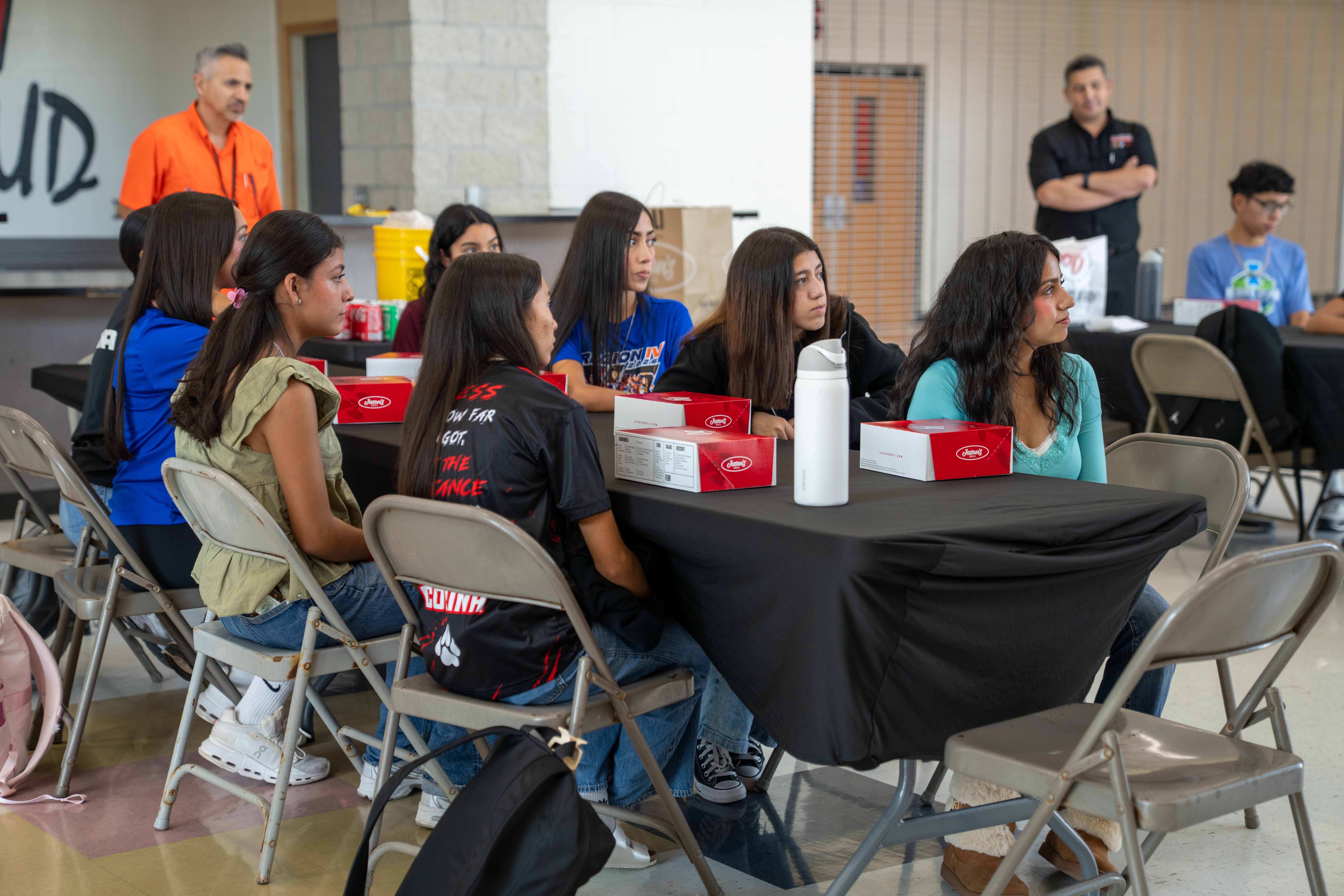 A group of students sits attentively at tables in a large hall, listening to a presentation.