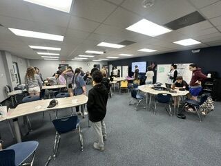A group of students gather in a brightly lit classroom, collaborating and talking around several desks.