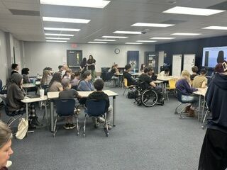 A group of students are seated at desks in a brightly lit classroom.
