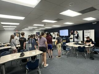 A group of students stands in a modern classroom, facing toward the front where a presentation screen is displayed.