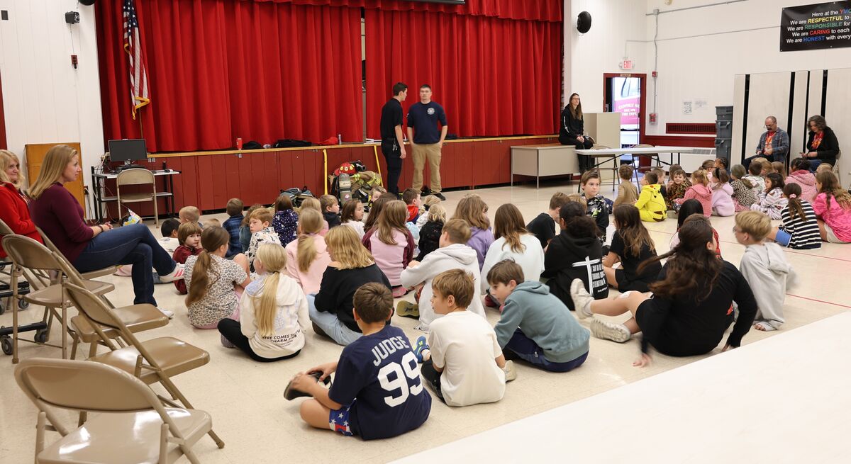 A group of children sits attentively on a gym floor, facing two firefighters standing near a stage with red curtains.