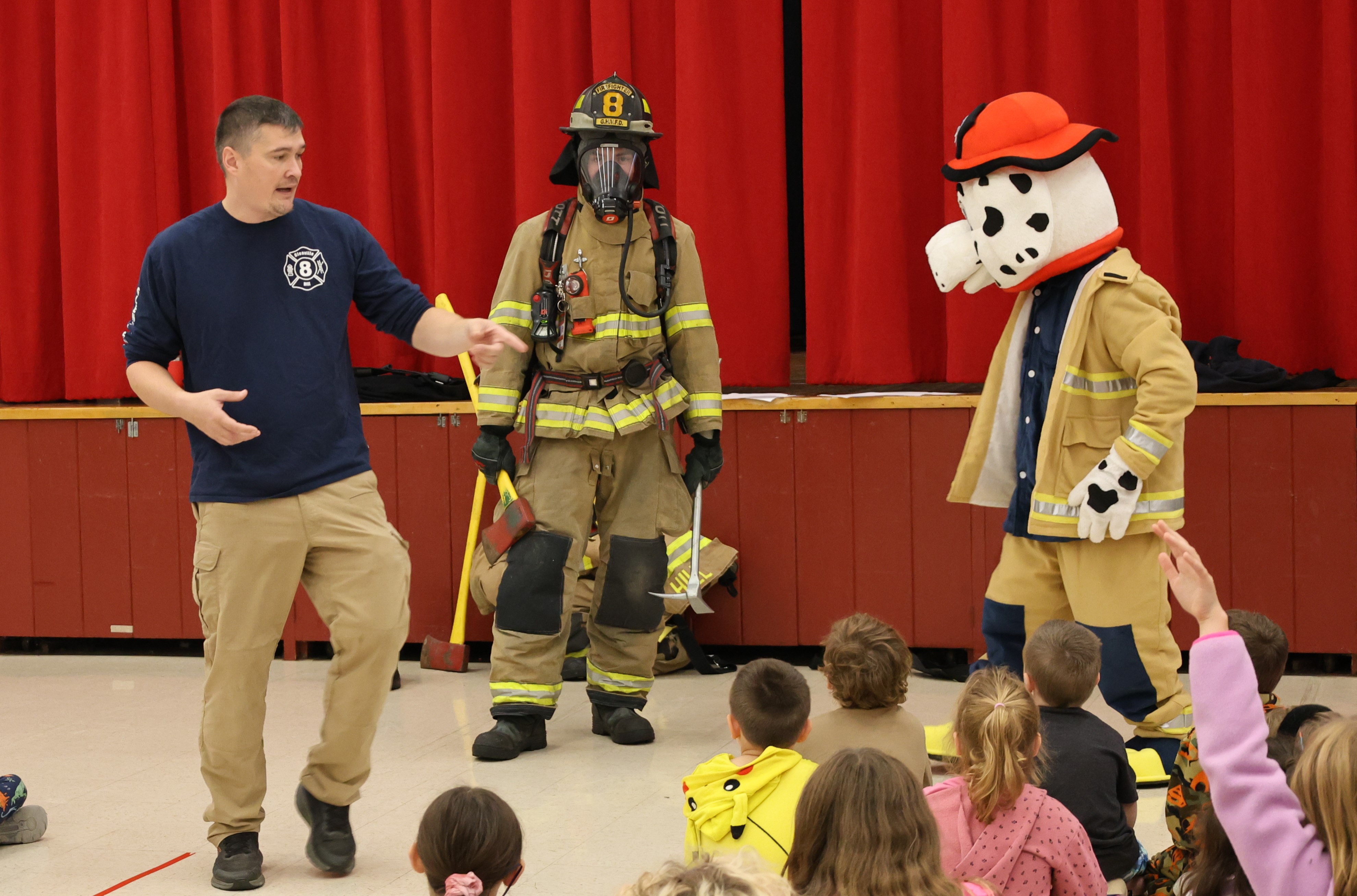 A firefighter in a blue shirt speaks to a group of children seated on the floor, while another person in full gear and a Dalmatian mascot stand behind him.