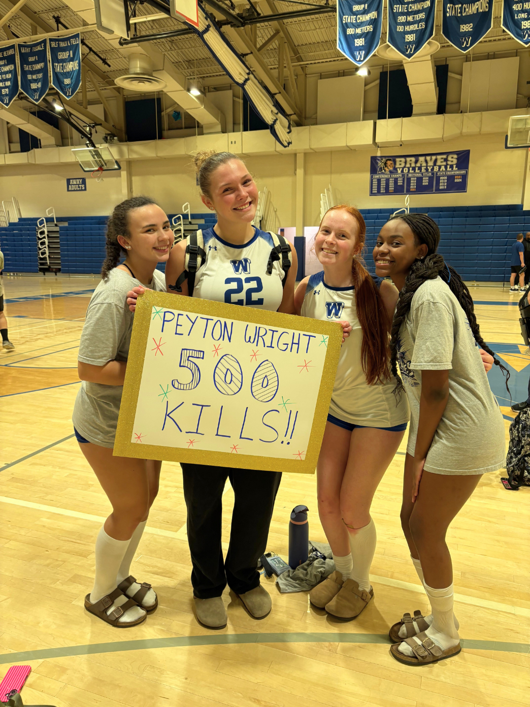 Four smiling volleyball players pose together in a gymnasium, holding a sign that reads, "Peyton Wright 500 kills!!".