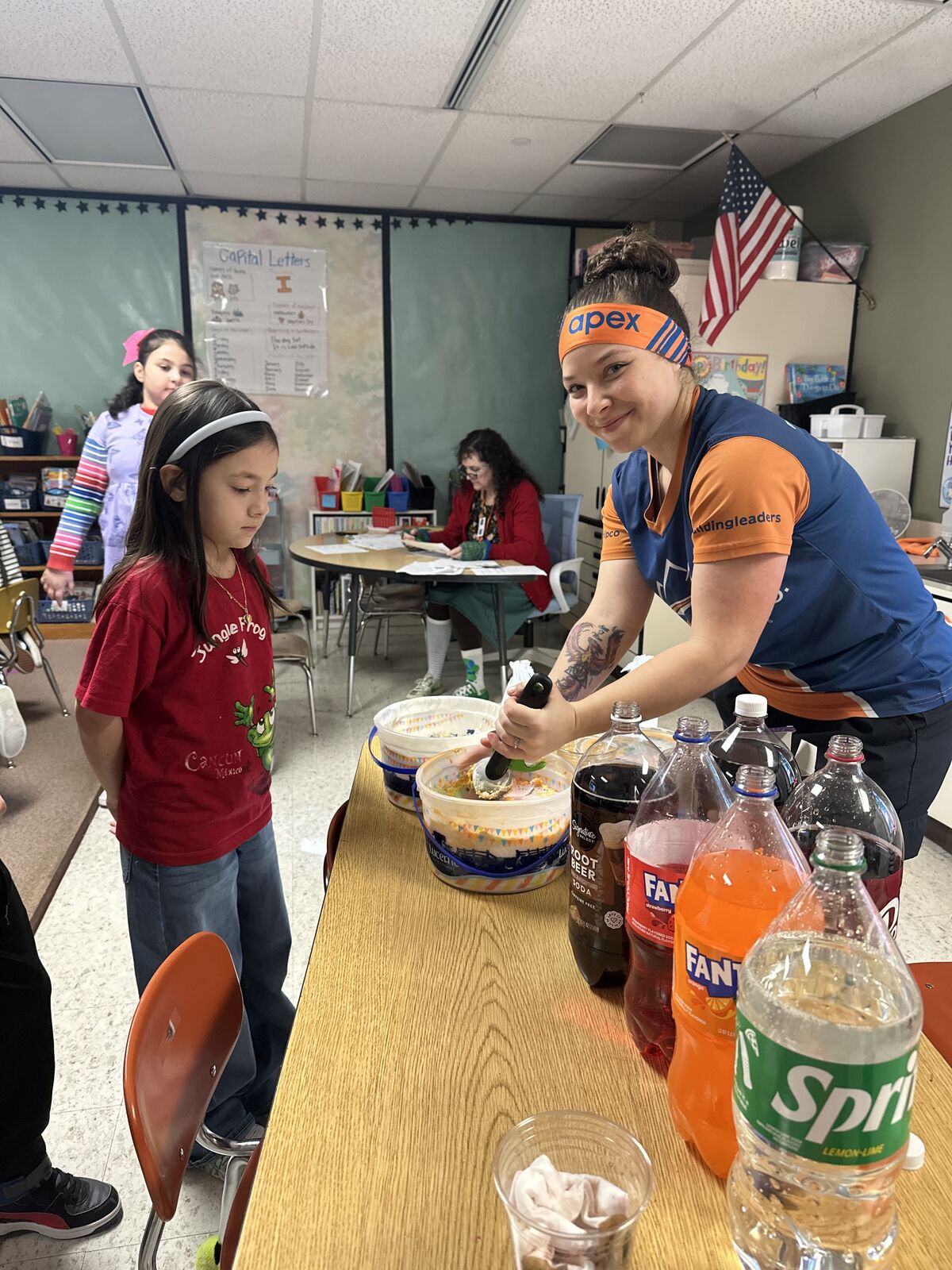 A teacher with an 'apex' headband pours drinks from soda bottles into buckets for students.