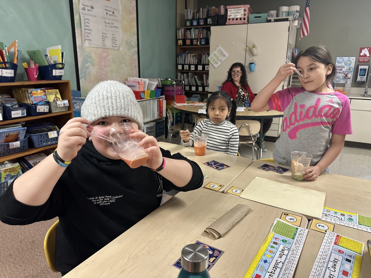 Three children are seated at desks in a classroom, each holding a cup with a drink and a utensil.