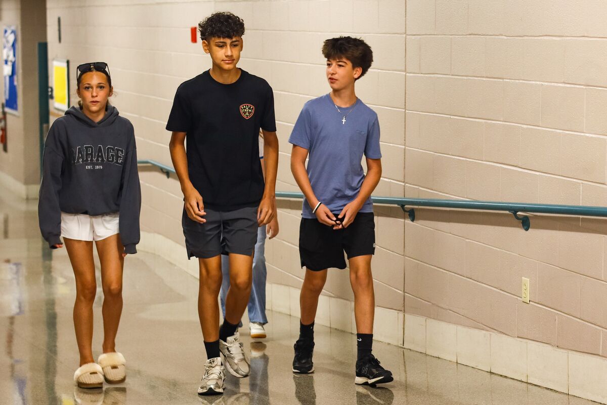 Three young people walk down a school hallway, with two boys in front and a girl behind.
