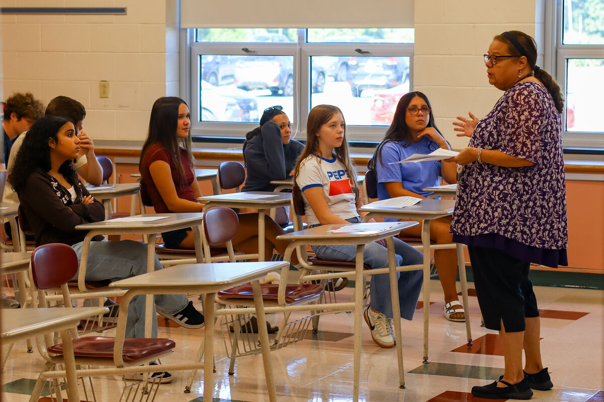 A teacher in a floral shirt addresses students seated at desks in a classroom.